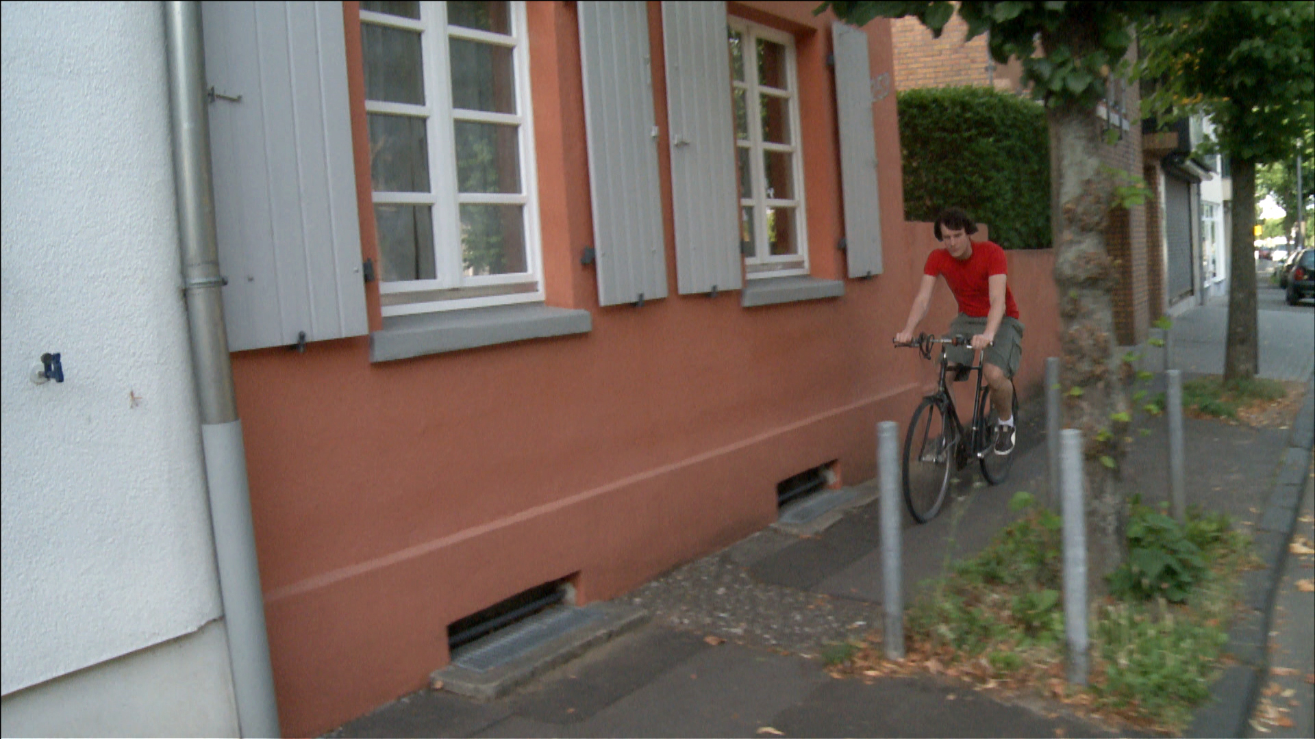 Ein Bürgersteig vor einer roten Hausfassade. Ein Radfahrer in rotem Shirt fährt durch eine Engstelle zwischen Fassade und einer Baumscheibe.