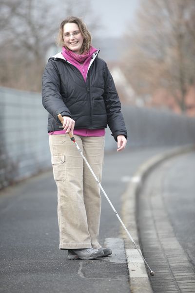 Eine junge Frau steht an der Bordsteinkante, ihr Langstock ertastet die Kante.