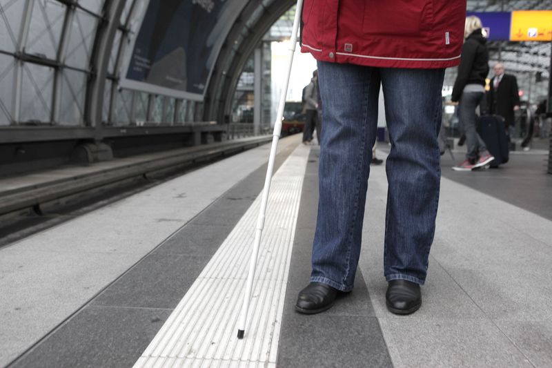 Eine Person mit einem weißen Langstock an einem Bahnsteig. Die Spitze des Stocks liegt auf dem Leitstreifen am Bahnsteigrand.