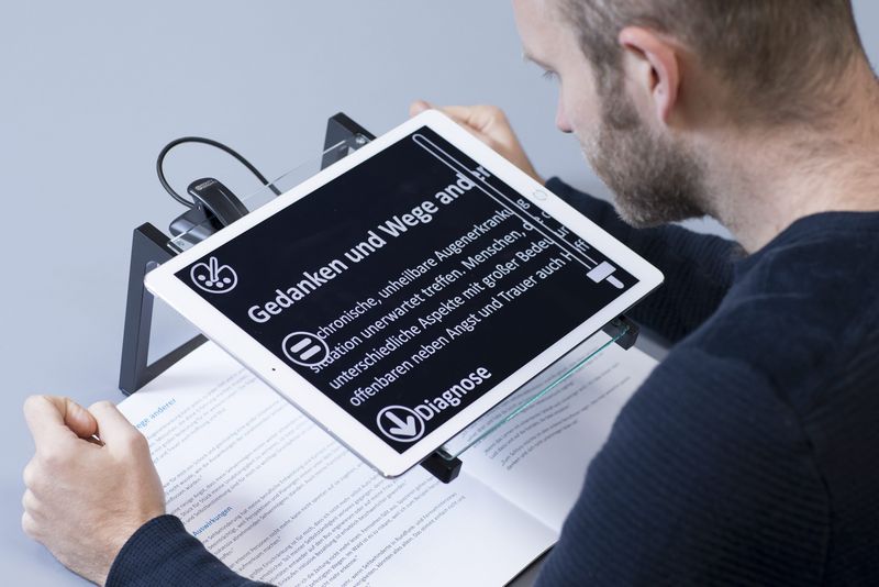 Ein Mann liest ein Buch mit Hilfe eines Tablets, das die Schrift vergrößert. Es ist auf ein Gestell mit etwas Abstand zum Buch montiert.